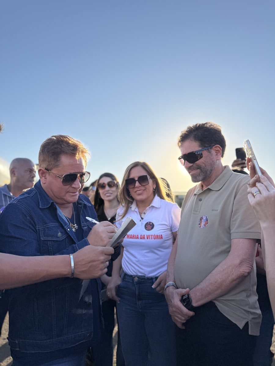 Bete de camiseta branca acompanha um apoiador dando autógrafo, ao ar livre no fim da tarde