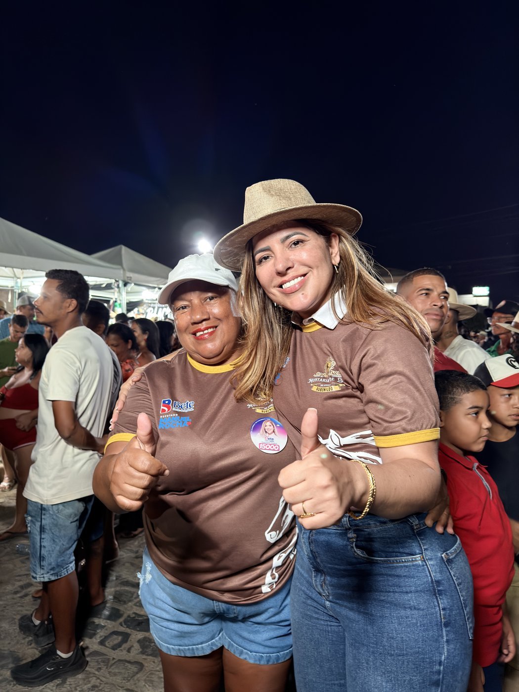 Bete de chapéu ao lado de uma apoiadora, ambas com o polegar erguido, em festa noturna